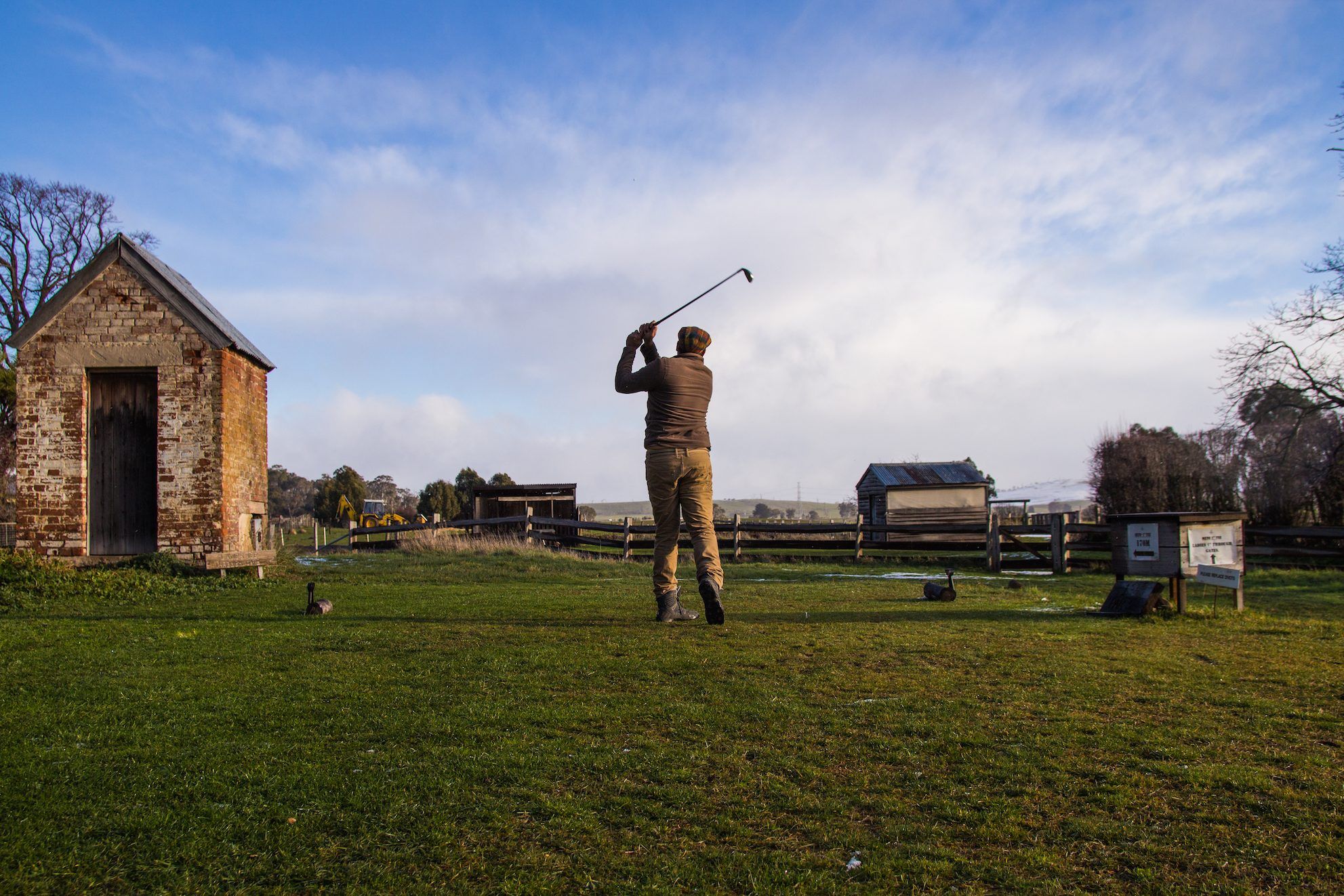 A golfer teeing off beside a historic stone hut at Ratho Farm